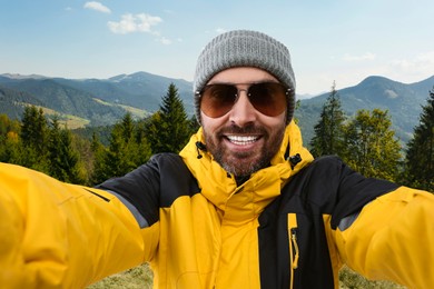 Image of Smiling man in hat and sunglasses taking selfie in mountains