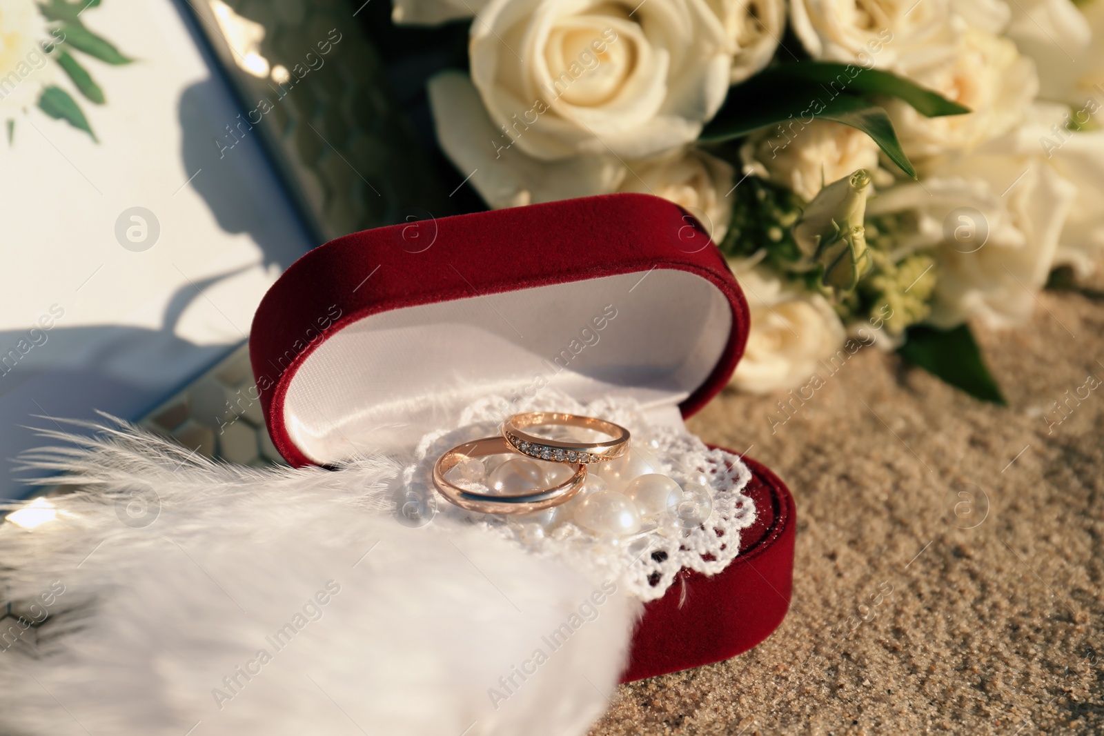 Photo of Red box with gold wedding rings and brooch on sandy beach, closeup