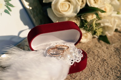 Photo of Red box with gold wedding rings and brooch on sandy beach, closeup