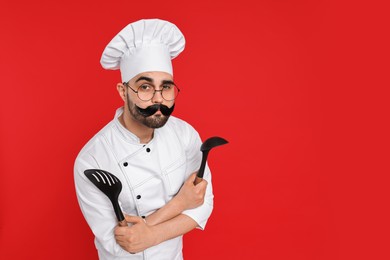 Photo of Professional chef with funny artificial moustache holding kitchen utensils on red background. Space for text