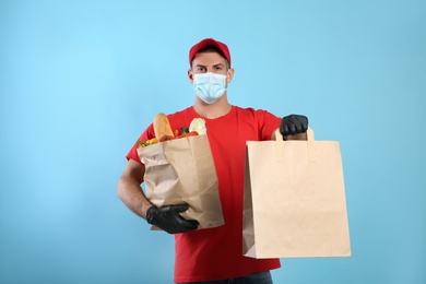 Photo of Courier in medical mask holding paper bags with takeaway food on light blue background. Delivery service during quarantine due to Covid-19 outbreak