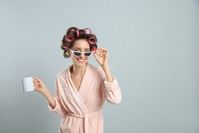 Photo of Beautiful young woman in bathrobe with hair curlers holding cup of drink on light grey background. Space for text