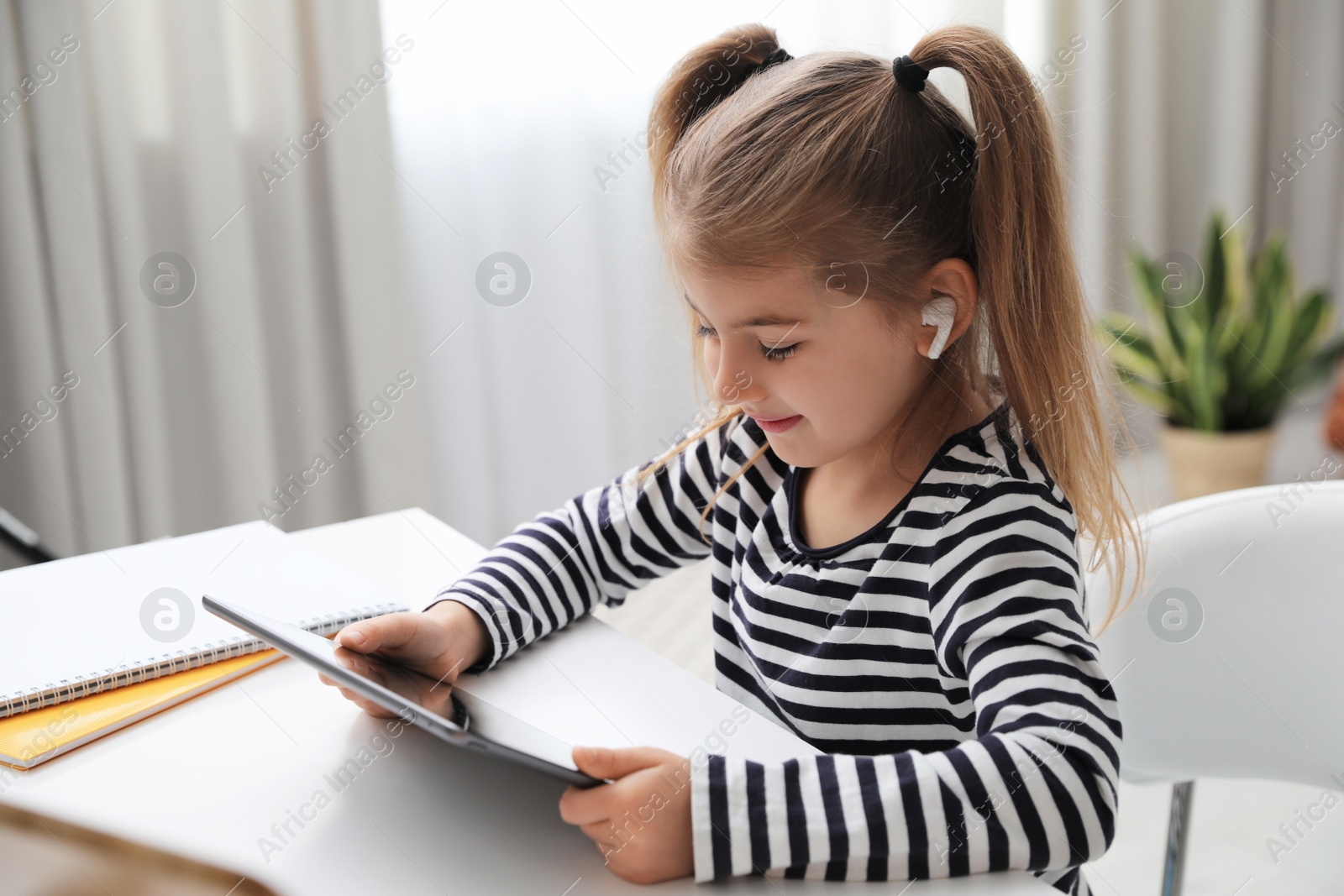 Photo of Adorable little girl doing homework with tablet and earphones at table indoors