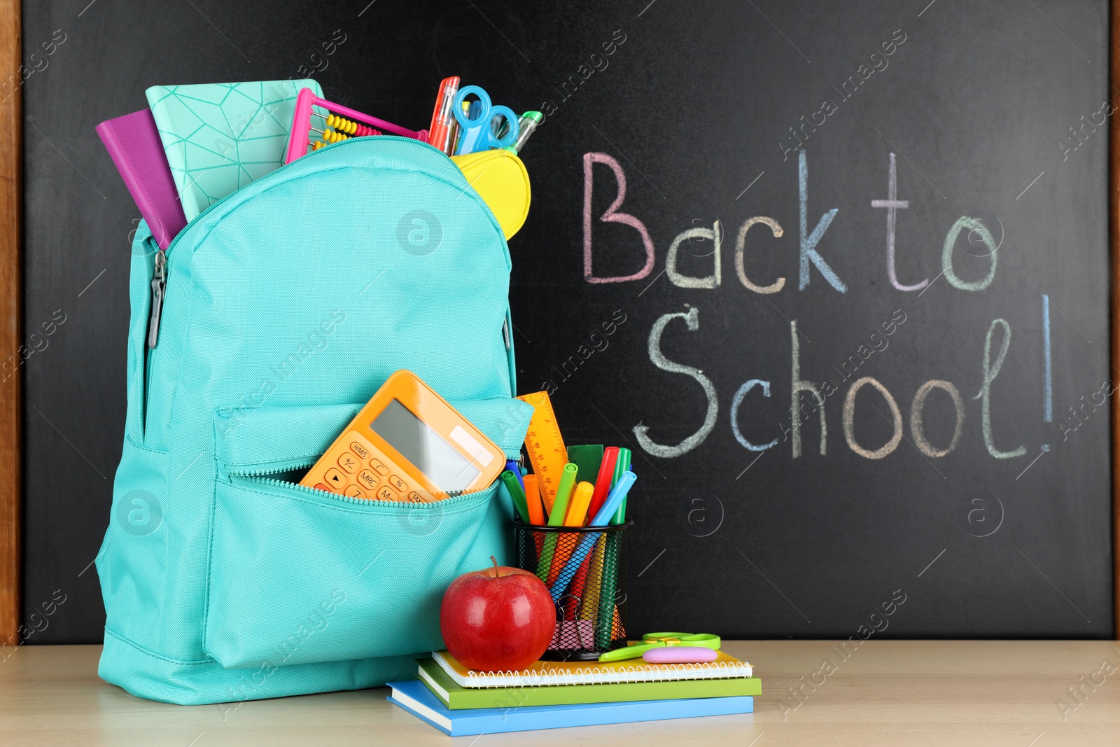 Photo of Bright backpack with school stationery on brown wooden table near black chalkboard. Back to School