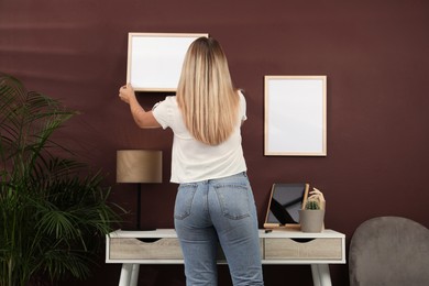 Woman hanging empty frame on brown wall indoors, back view. Mockup for design