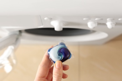 Photo of Woman holding laundry detergent capsule near washing machine indoors, top view