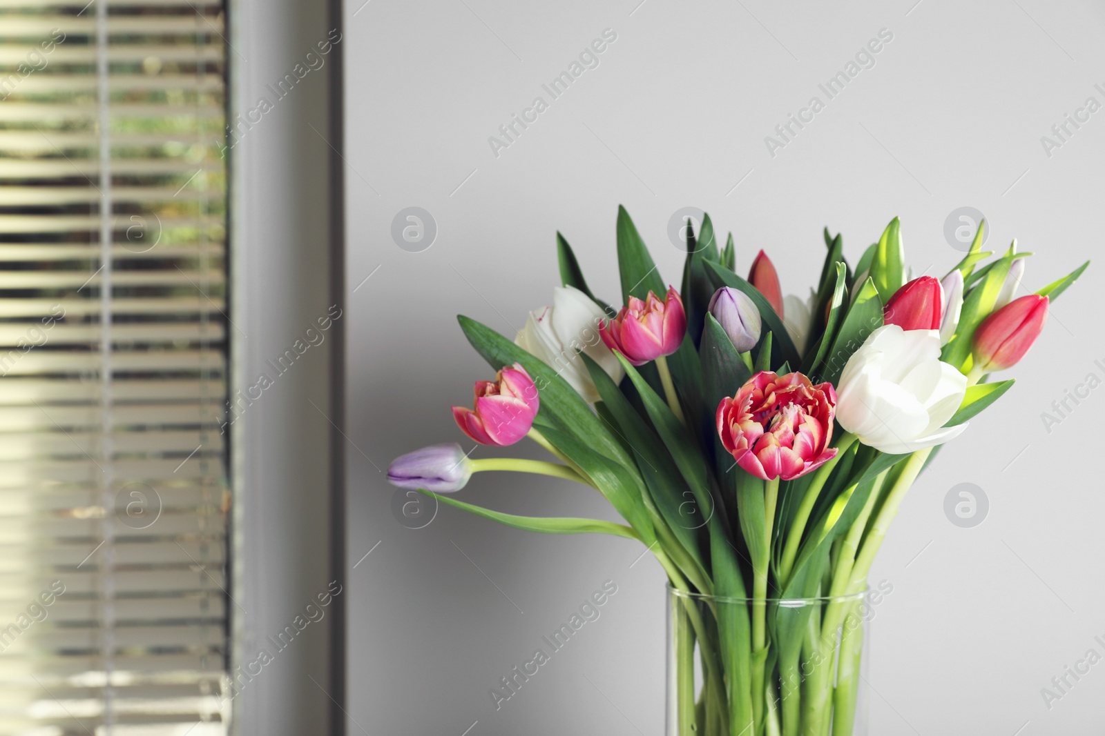 Photo of Beautiful bouquet of colorful tulips in glass vase near beige wall at home