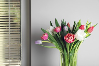 Photo of Beautiful bouquet of colorful tulips in glass vase near beige wall at home