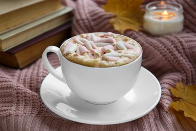 Photo of Cup of tasty hot drink with marshmallows, books, leaves and burning candle on blanket, closeup. Autumn coziness