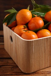 Photo of Fresh tangerines with green leaves in crate on wooden table, closeup