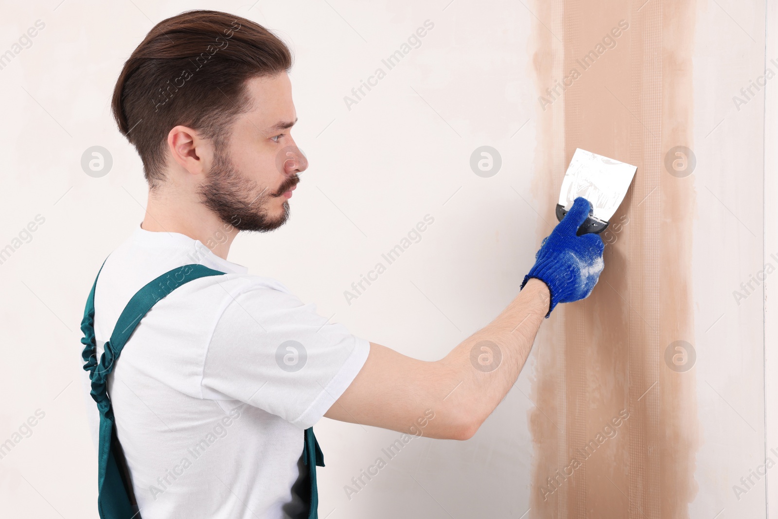 Photo of Worker in uniform plastering wall with putty knife indoors