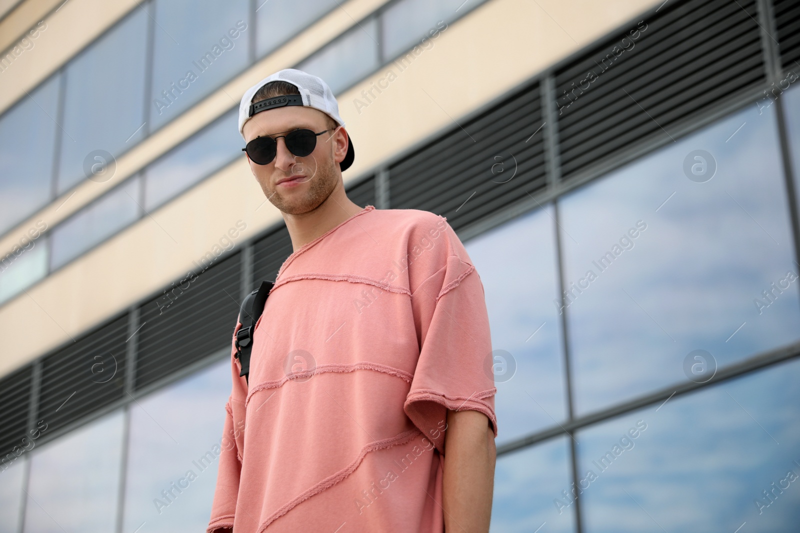 Photo of Handsome young man with stylish sunglasses and backpack near building outdoors, low angle view