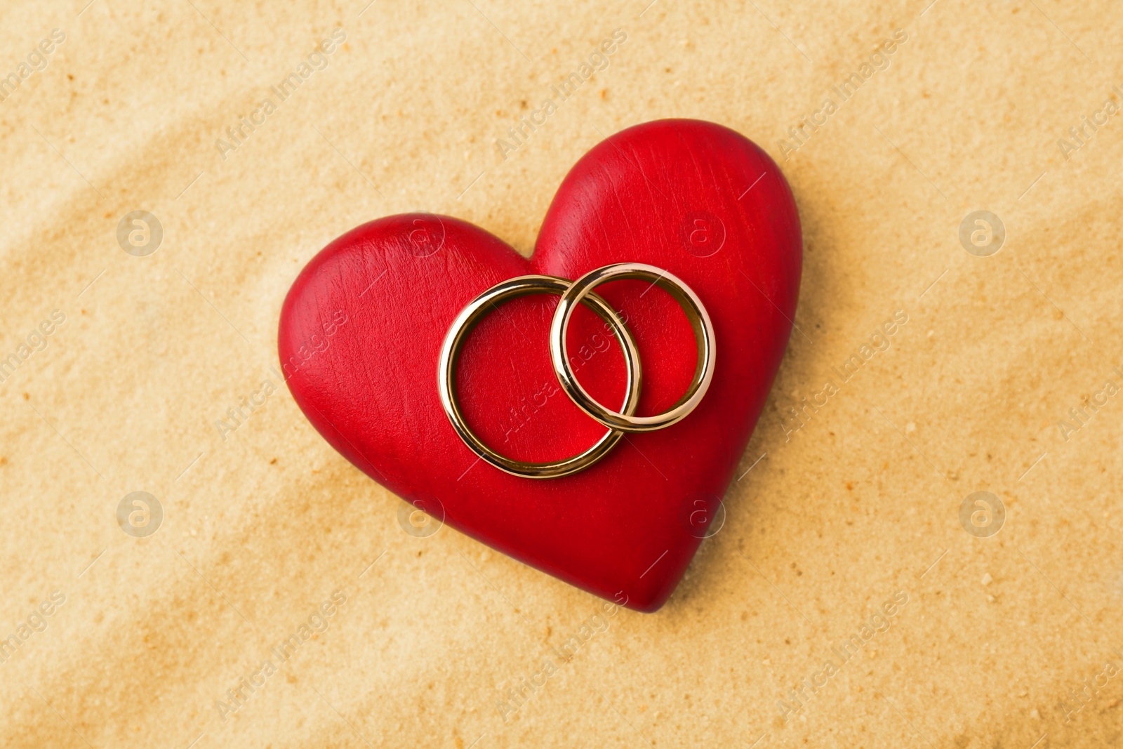 Photo of Honeymoon concept. Two golden rings and red wooden heart on sand, top view