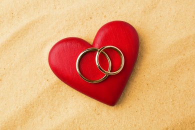 Photo of Honeymoon concept. Two golden rings and red wooden heart on sand, top view