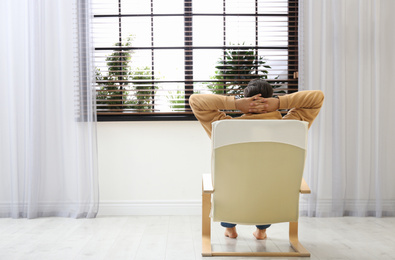 Photo of Man relaxing in armchair near window at home, back view. Space for text