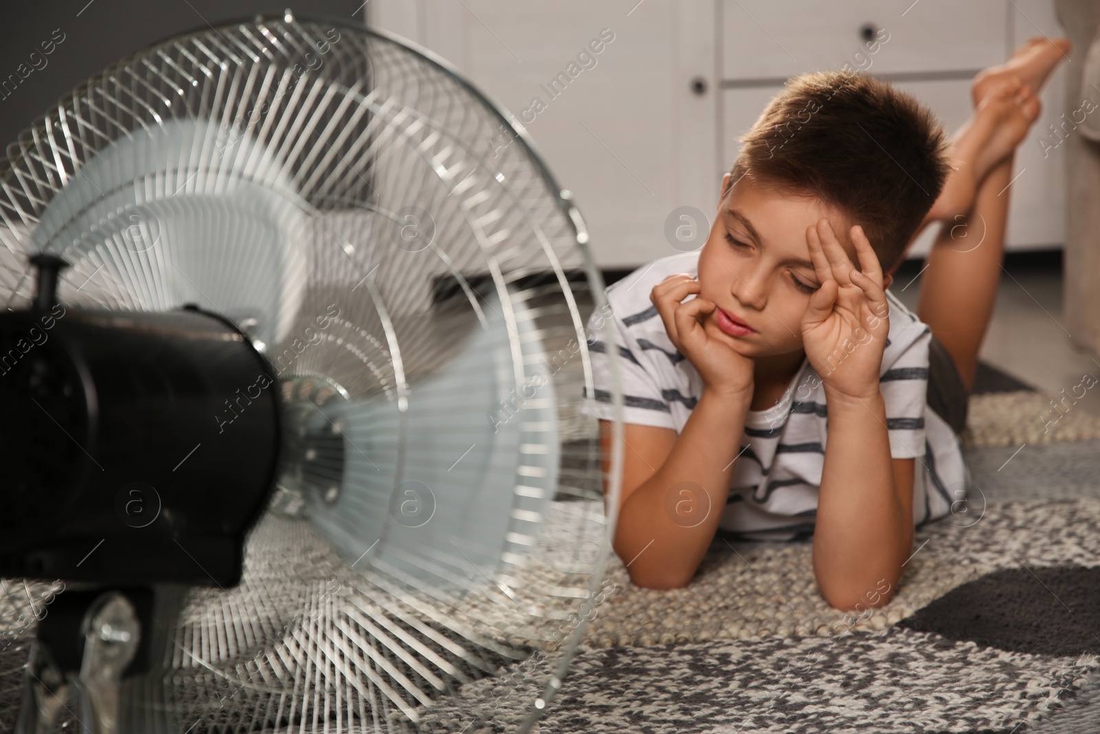 Photo of Little boy suffering from heat in front of fan at home. Summer season