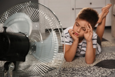 Photo of Little boy suffering from heat in front of fan at home. Summer season