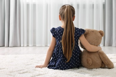 Photo of Cute little girl with teddy bear at home, back view. Space for text