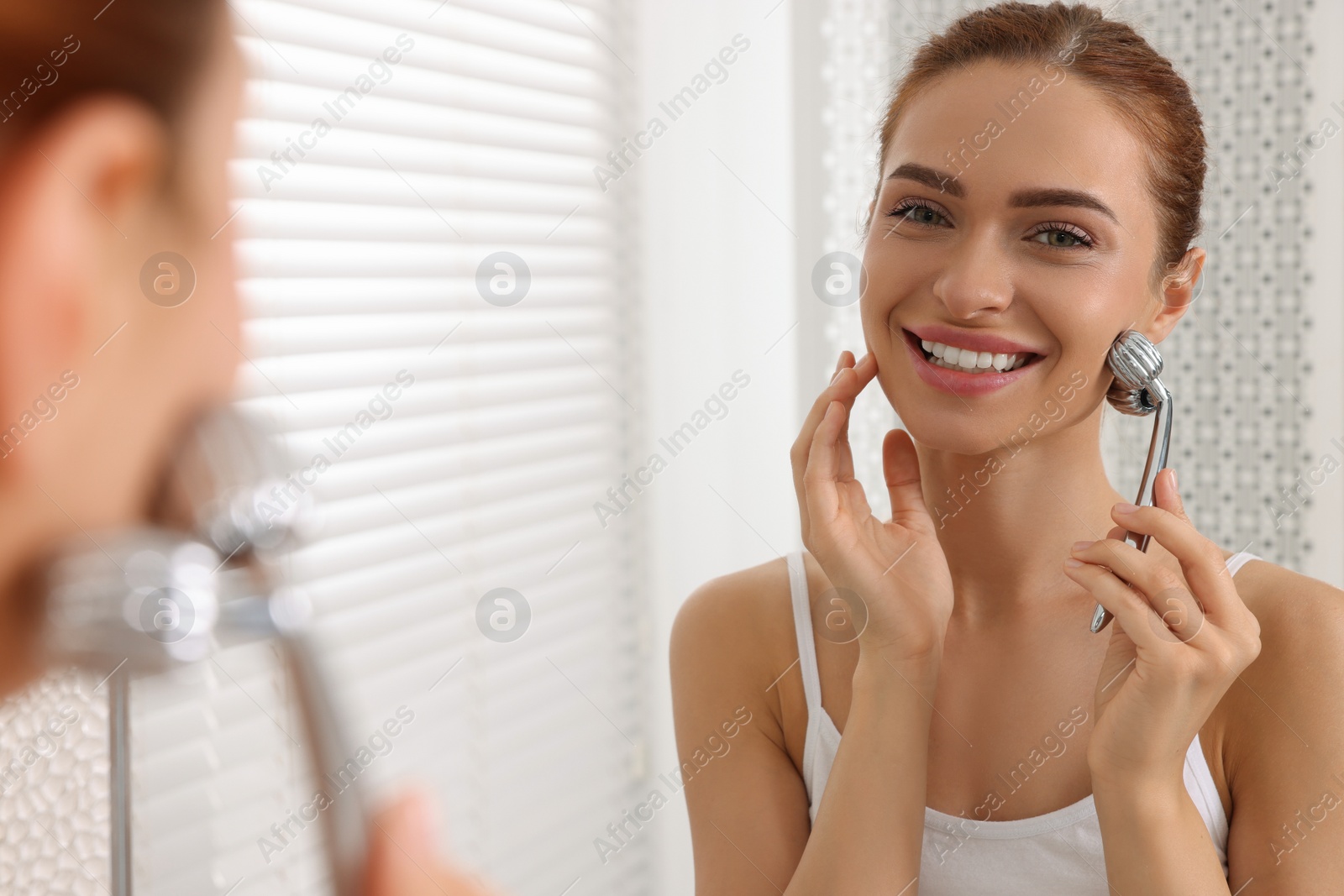 Photo of Young woman massaging her face with metal roller near mirror in bathroom