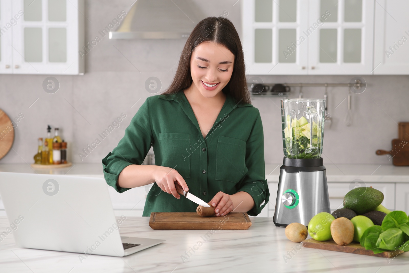 Photo of Young woman cutting kiwi for smoothie at white table in kitchen