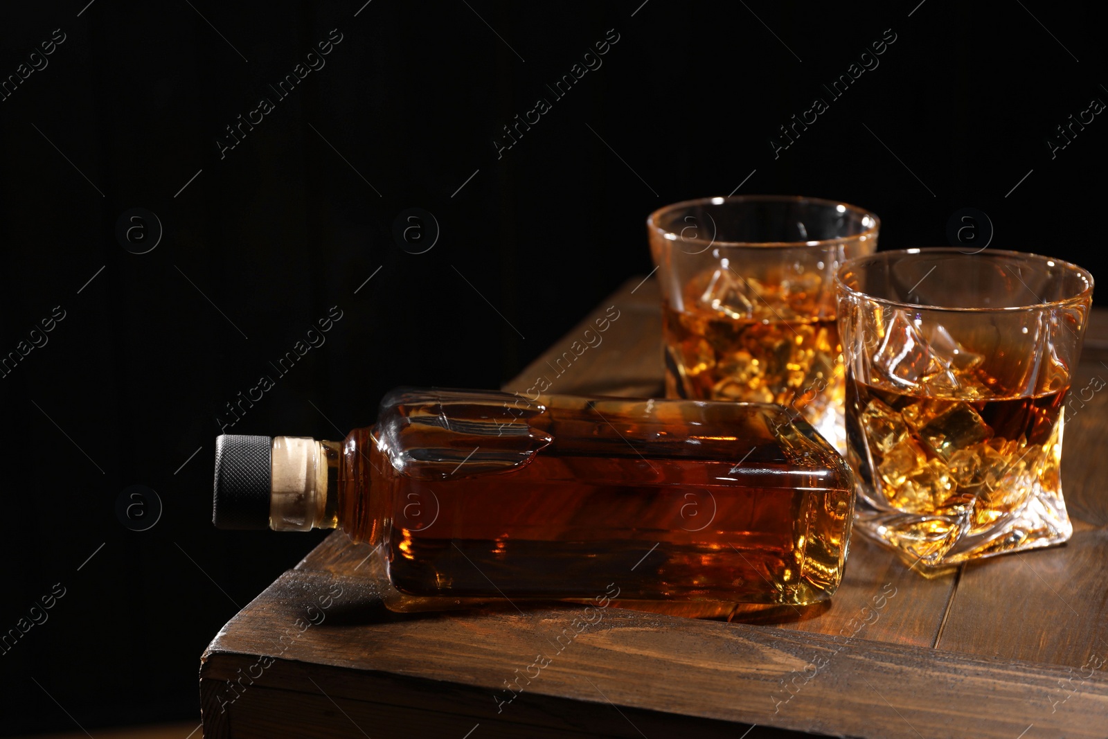 Photo of Whiskey with ice cubes in glasses and bottle on wooden crate against black background, closeup