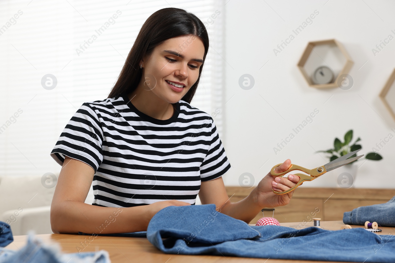 Photo of Young woman with jeans and scissors at wooden table indoors