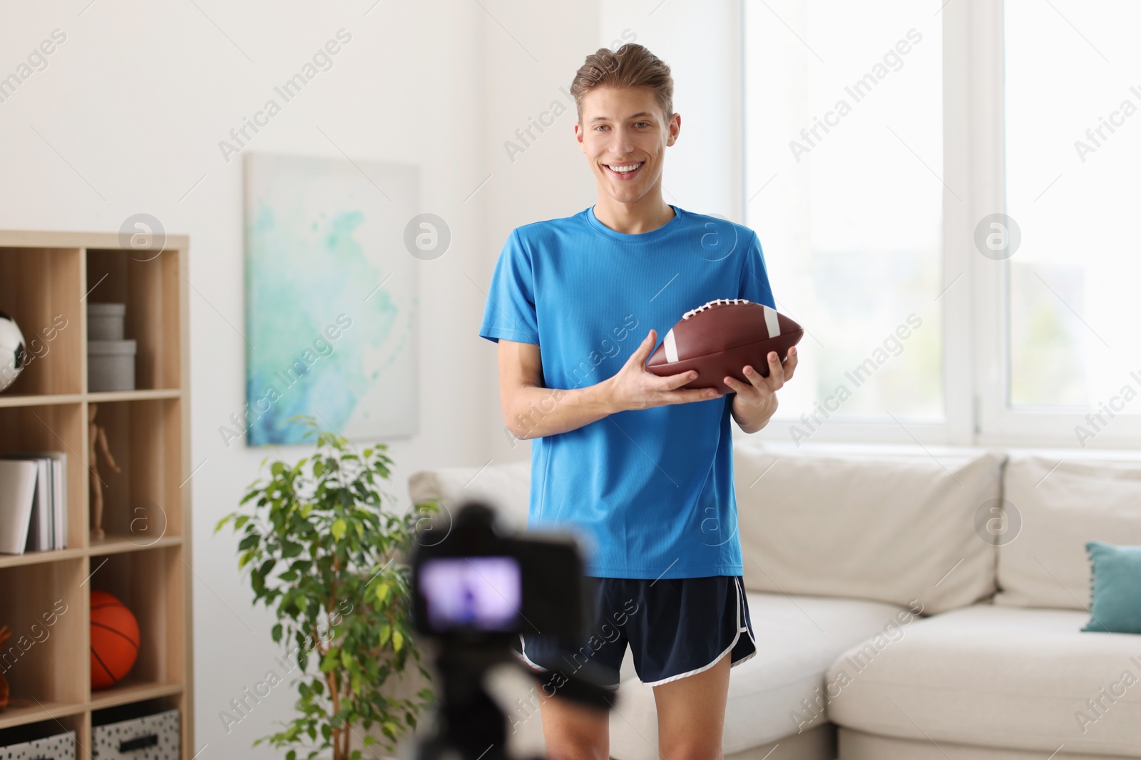 Photo of Smiling sports blogger holding american football ball while recording fitness lesson with camera at home