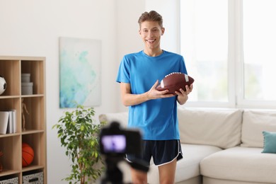 Smiling sports blogger holding american football ball while recording fitness lesson with camera at home