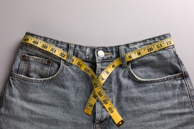 Jeans with measuring tape on light grey background, top view. Weight loss concept