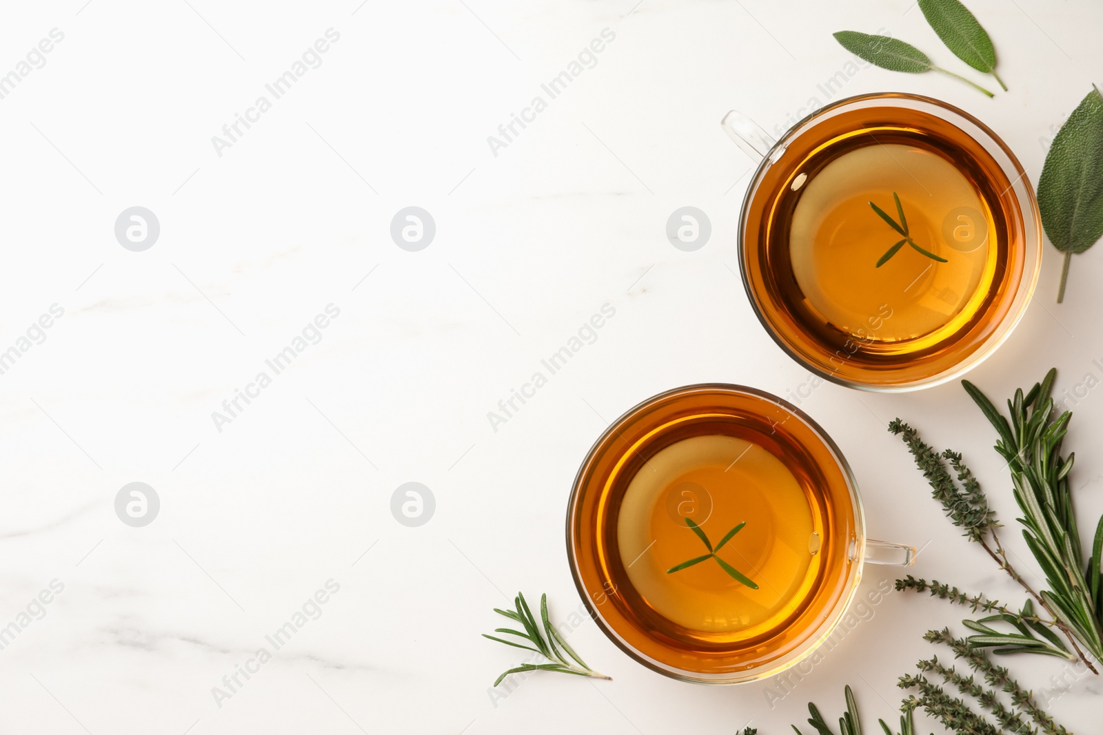 Photo of Cups of aromatic herbal tea with thyme, rosemary and sage on white marble table, flat lay. Space for text