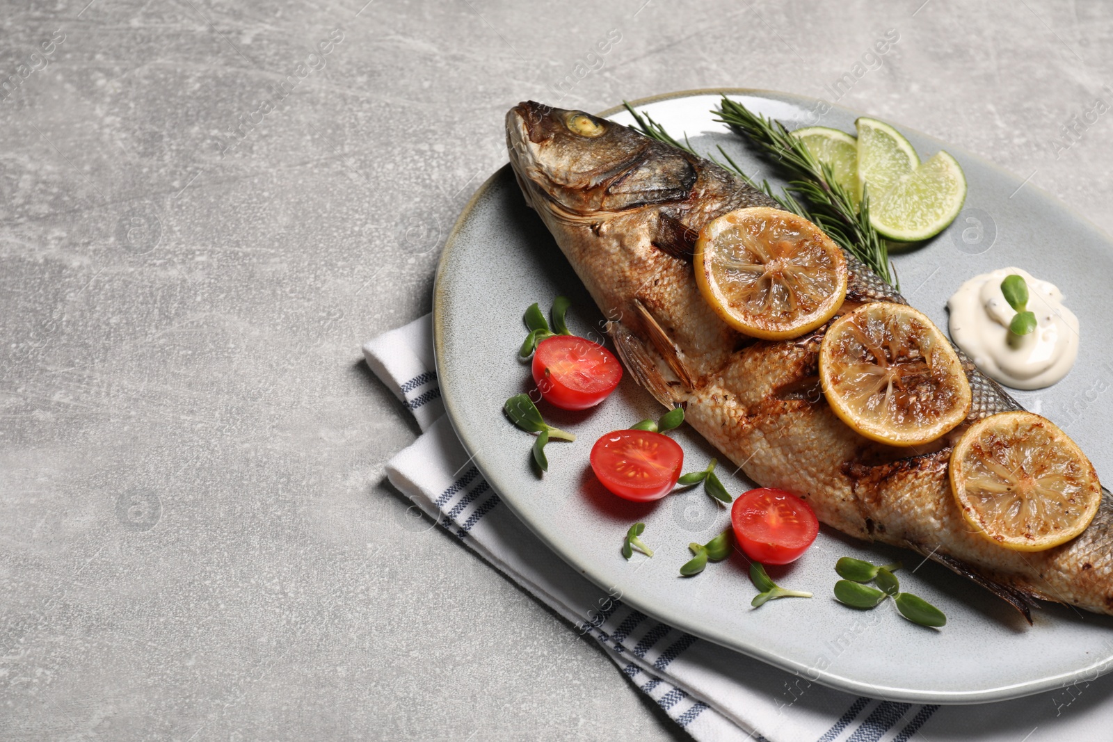Photo of Delicious baked sea bass fish served with lemon, tomatoes and sauce on light grey table. Space for text