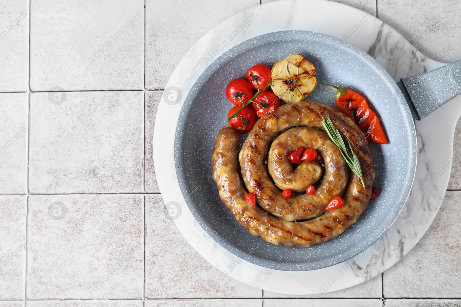 Photo of Delicious homemade sausage with garlic, tomatoes, rosemary and chili in frying pan on light tiled table, top view. Space for text