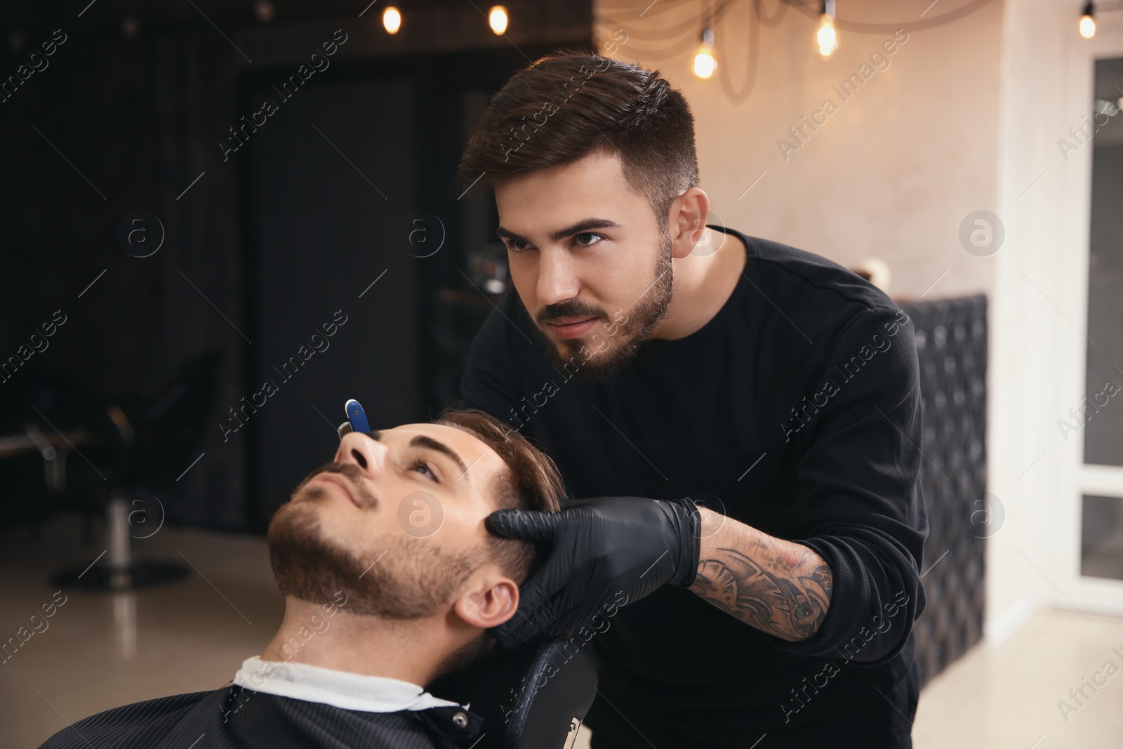 Photo of Professional hairdresser shaving client with straight razor in barbershop