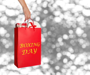 Image of Woman holding red shopping bag with text Boxing Day full of gifts on blurred silver background, closeup. Space for text