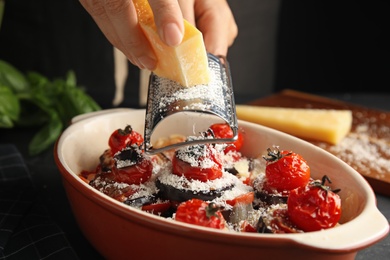 Photo of Woman grating cheese onto baked eggplant with tomatoes at table, closeup