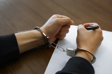Photo of Criminal in handcuffs writing confession at desk, closeup