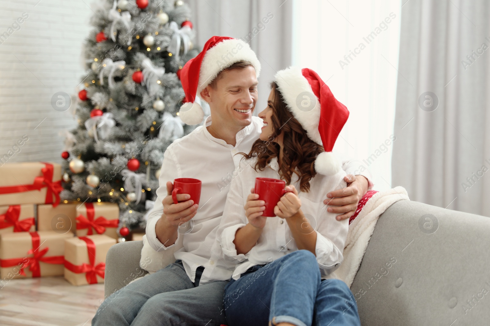 Image of Happy couple in Santa hats with cups of hot drink near Christmas tree at home