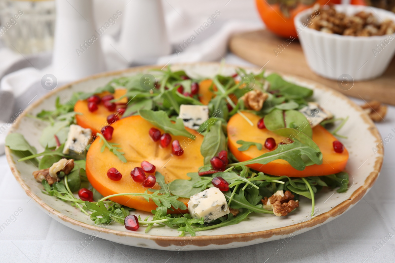 Photo of Tasty salad with persimmon, blue cheese, pomegranate and walnuts served on white tiled table, closeup