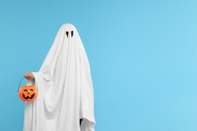 Photo of Woman in white ghost costume holding pumpkin bucket on light blue background, space for text. Halloween celebration