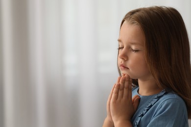 Photo of Cute little girl with hands clasped together praying indoors. Space for text