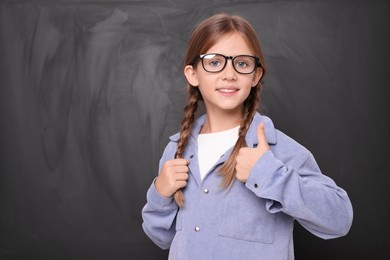 Photo of Cute schoolgirl in glasses showing thumb up near blackboard. Space for text