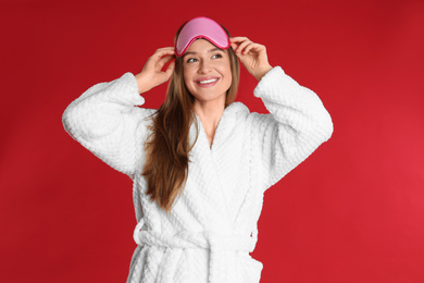 Young woman in bathrobe and eye sleeping mask on red background