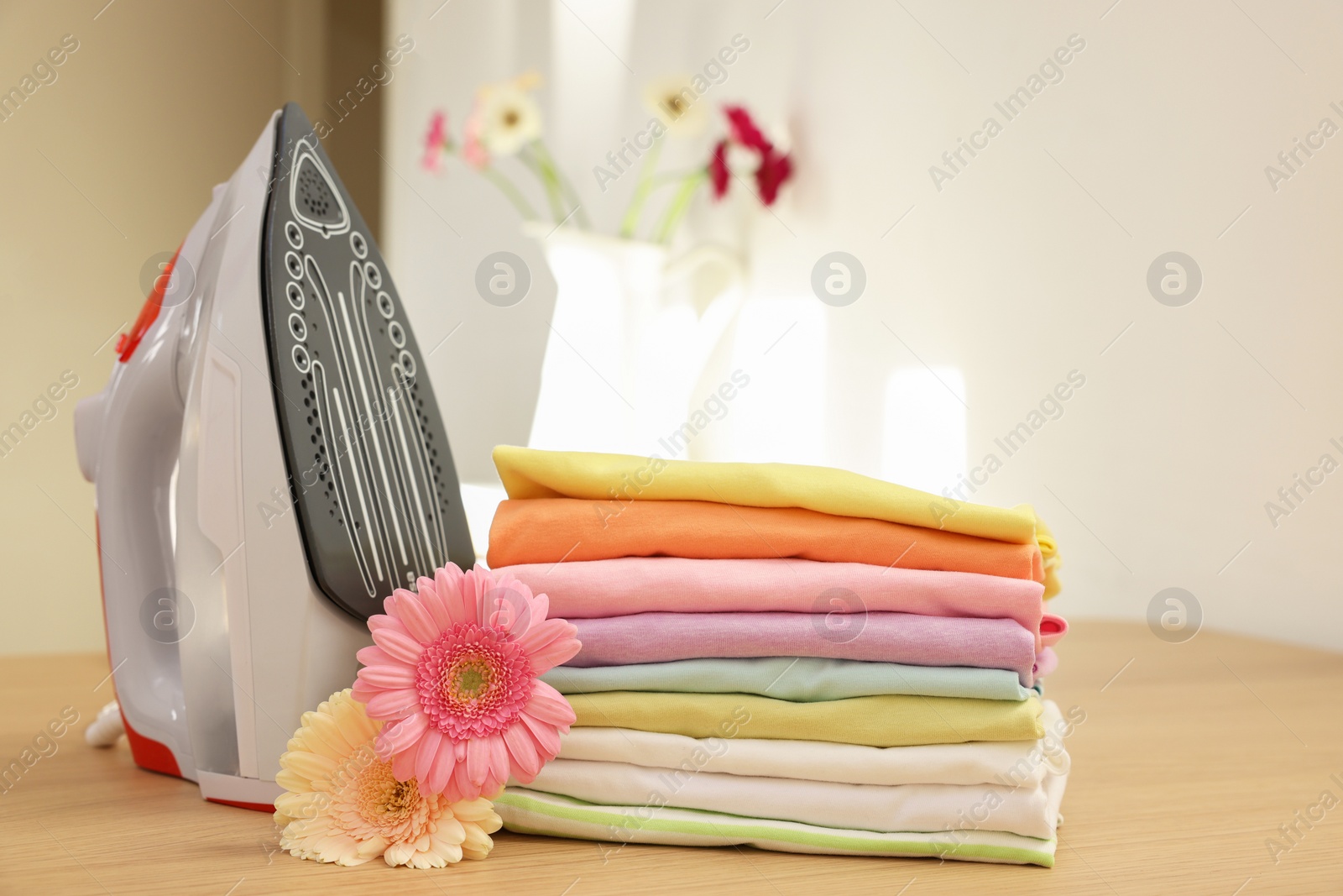 Photo of Stack of clean clothes, modern iron and flowers on wooden table