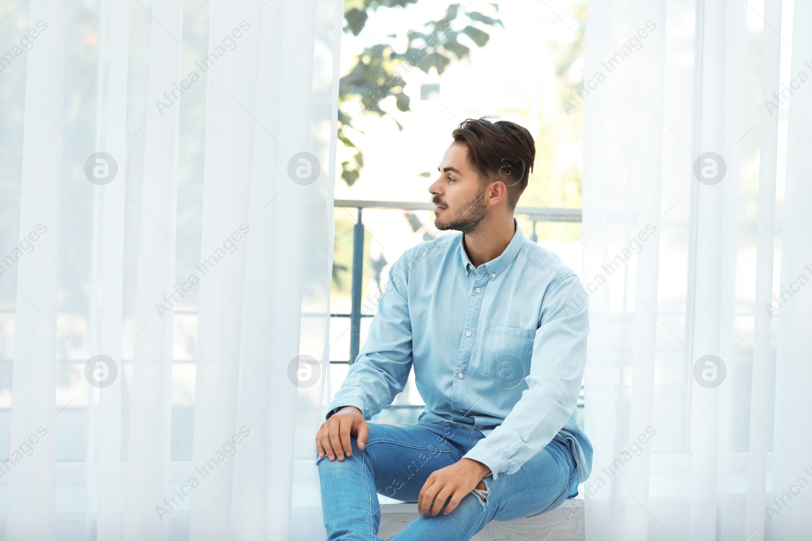 Photo of Young man sitting near window with beautiful curtains at home