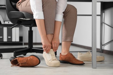 Photo of Woman taking off uncomfortable shoes and putting on sneakers in office, closeup