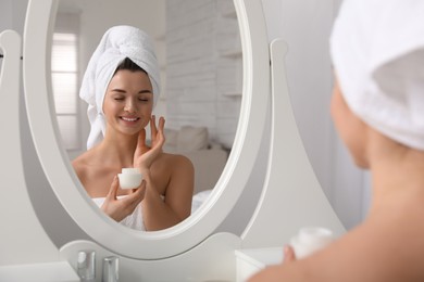 Photo of Beautiful young woman with hair wrapped in towel applying cream near mirror indoors