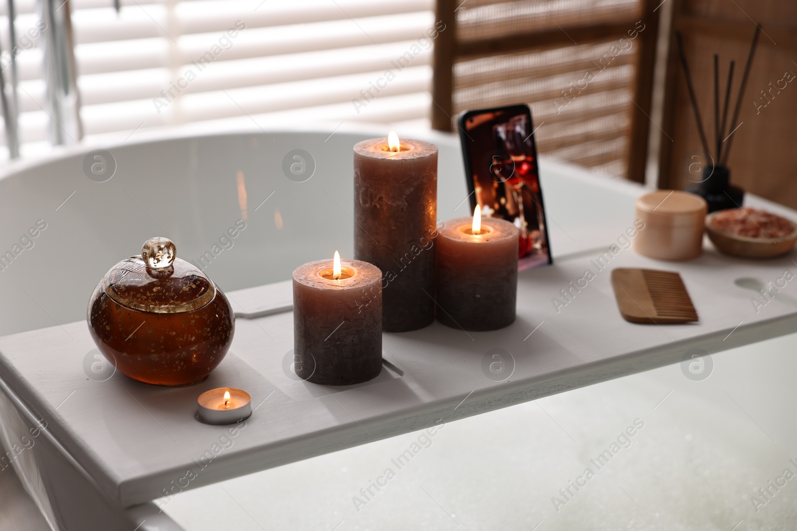 Photo of White wooden tray with burning candles and beauty products on bathtub in bathroom