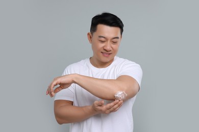 Handsome man applying body cream onto his elbow on light grey background