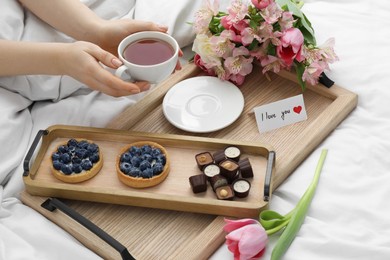 Photo of Tasty breakfast served in bed. Woman with tea, desserts, flowers and I Love You card at home, closeup