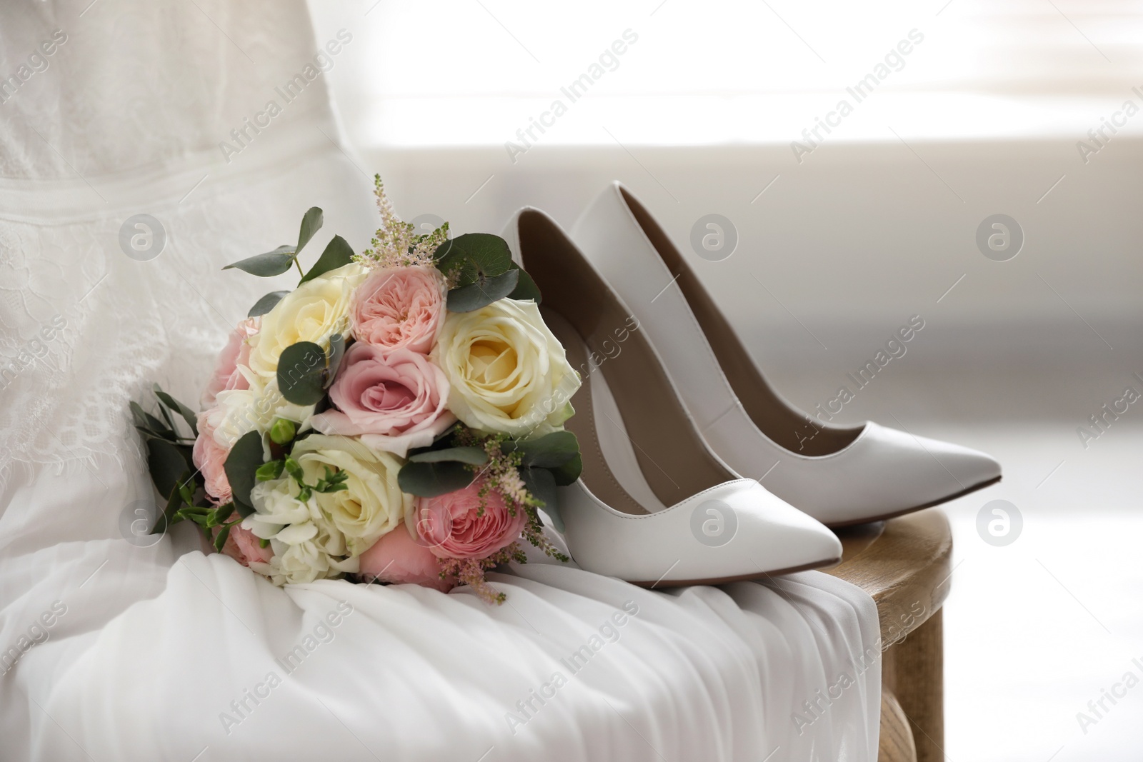 Photo of White high heel shoes, flowers and wedding dress on wooden chair indoors